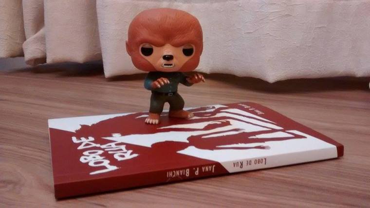 O livro "Lobo de Rua" está deitado em cima do piso de chão de madeira. Sobre ele, está um bonequinho de plástico em forma de lobisomem.