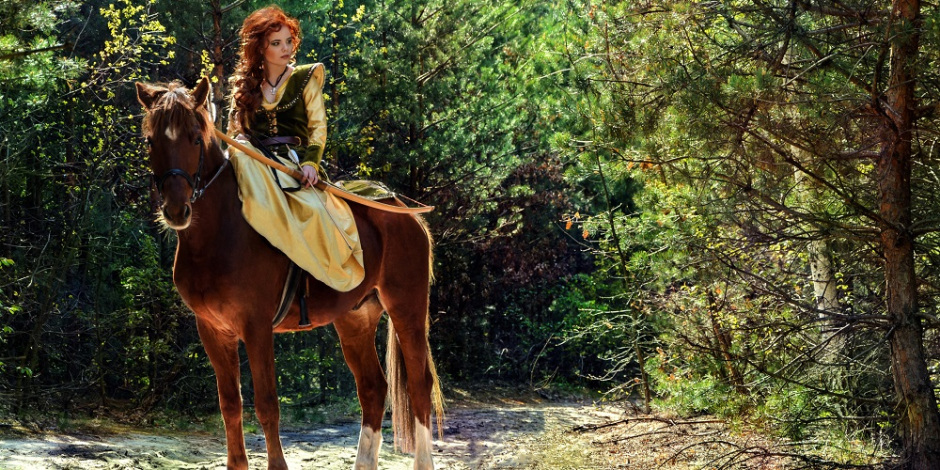 Uma mulher de roupa medieval está montada em um cavalo em uma floresta. Ela segura um arco e flechas.