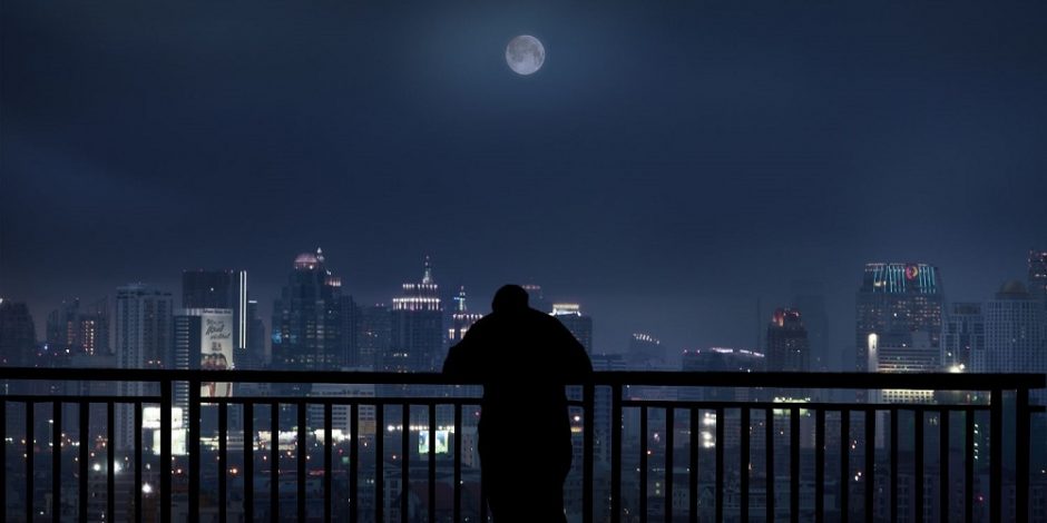 Um homem está na varanda de um apartamento no meio da noite. Ele observa a cidade abaixo enquanto a lua ilumina o céu.