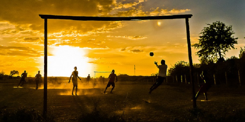 Fotografia de um campo de várzea. Algumas pessoas jogam futebol ao entardecer em um campo de terra e grama. O observador está posicionado atrás de um dos gols feitos de pedaços de madeira, onde é possível ver o goleiro pulando para defender uma bola.