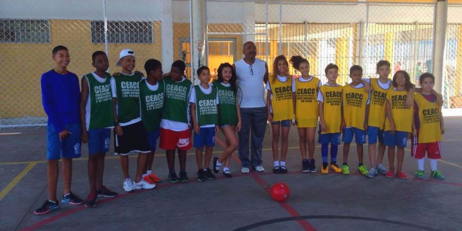 Zezé, o goleiro da Cidade de Deus, está no meio da foto. De cada lado seu, estão crianças enfileiradas. Do lado direito da foto, estão crianças usando colete amarelo do projeto CEAC. Do lado esquerdo, estão crianças usando colete verde do projeto CEAC. Todos olham para frente e estão no meio da quadra de futebol de uma escola. Na frente, está uma bola vermelha.