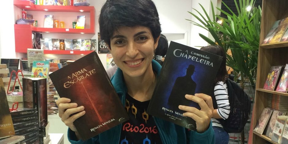 A escritora Renata Ventura está em estande na Bienal do Livro. Ela segura em cada uma das mãos um de seus livros. Na mão direita está "A Arma Escarlate". Na mão esquerda está "A Comissão Chapeleira". Ao seu redor, estão vários livros.